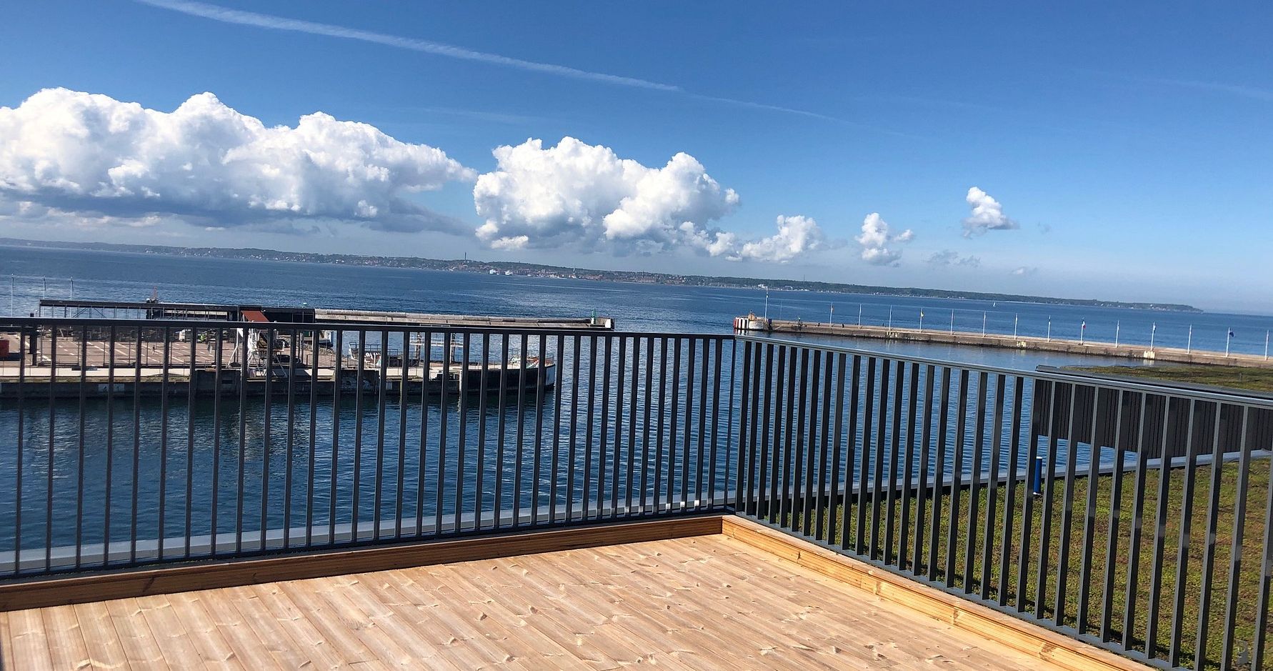 Vy från en träbalkong med räcke mot en hamn och havet, under en klarblå himmel med moln