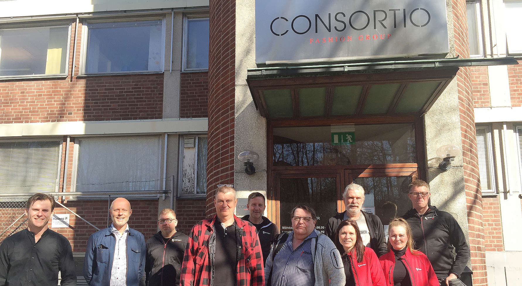 Grupp av människor framför en byggnad med en skylt som säger "Consortio". Byggnaden har röda tegelväggar och stora fönster
