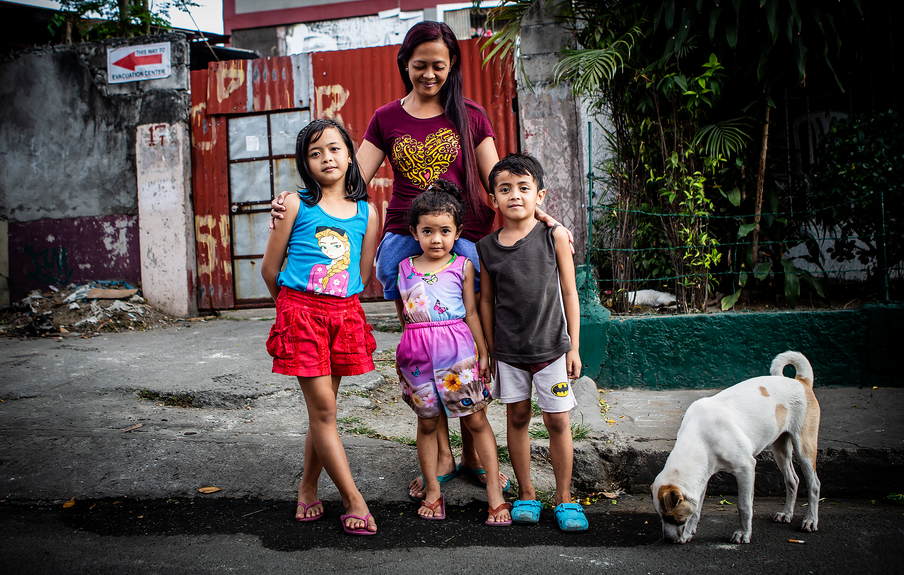 En kvinna står utomhus med fyra barn klädda i färgglada kläder, bredvid en vit hund på en gata framför en grind och vegetation