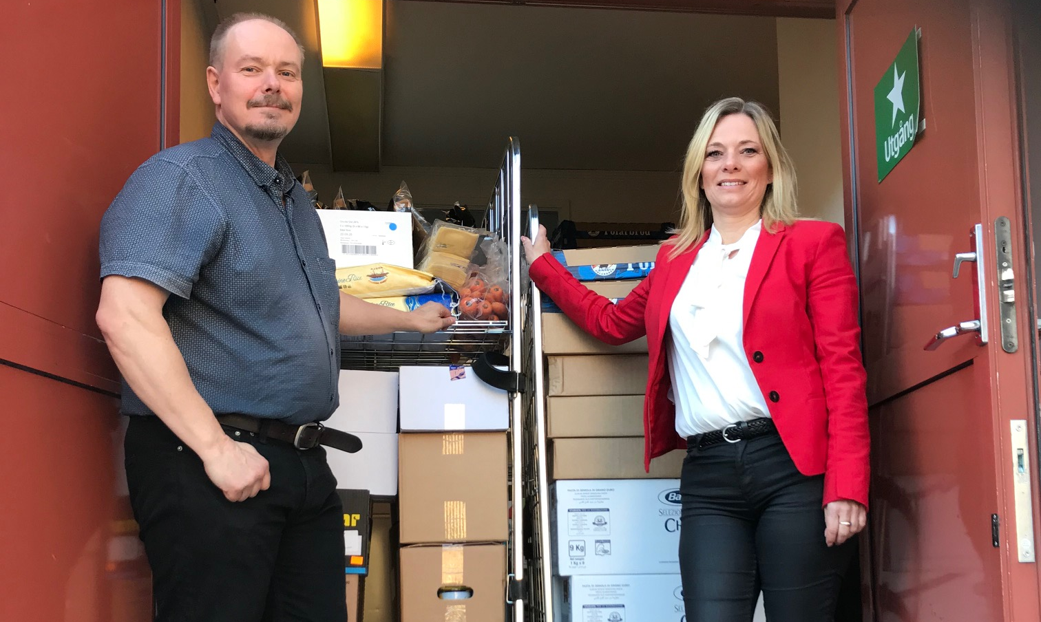 Två personer står vid en dörröppning fylld med lådor, påsar och andra leveranser; en håller i en stång till en vagn