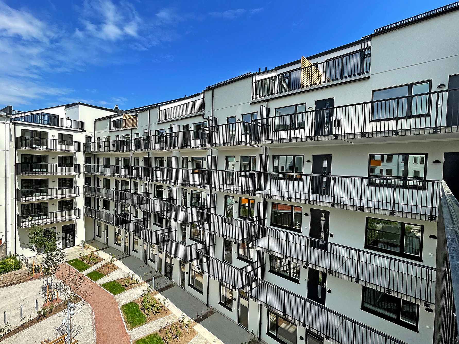 Bostadskomplex med modern arkitektur, flera våningar och balkonger, beläget vid en innergård med gångvägar och planteringar