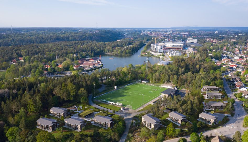 Luftvy över ett grönområde med en fotbollsplan, omgivande bostäder och en sjö i bakgrunden