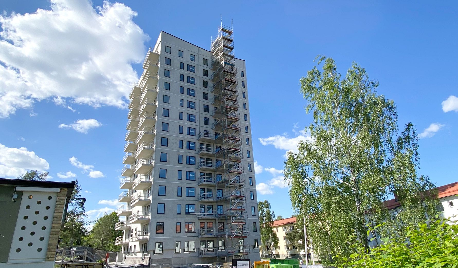 Höghus under konstruktion med byggnadsställningar, omgiven av träd och grönska under en klarblå himmel