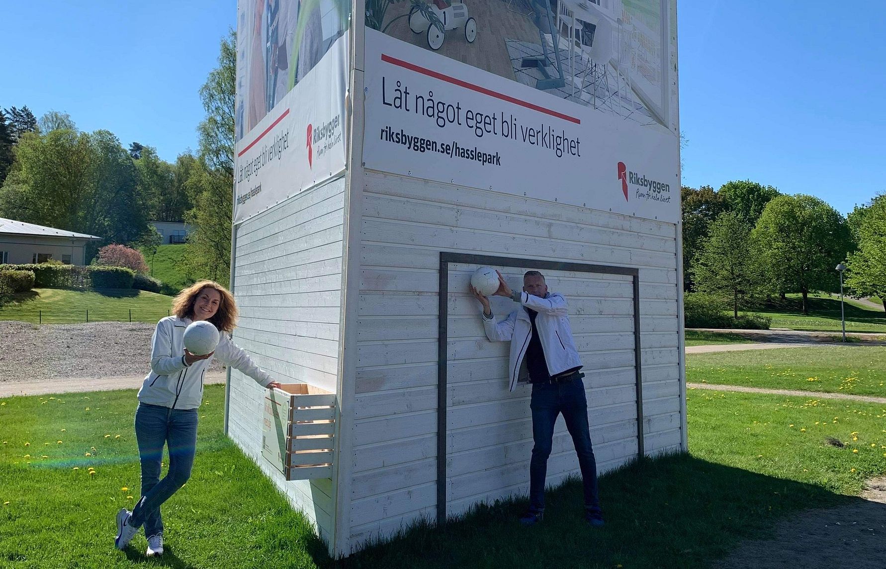 Personer står vid en vit byggnad med skyltar för Riksbyggen på en grön gräsmatta, en person håller en vit hjälm
