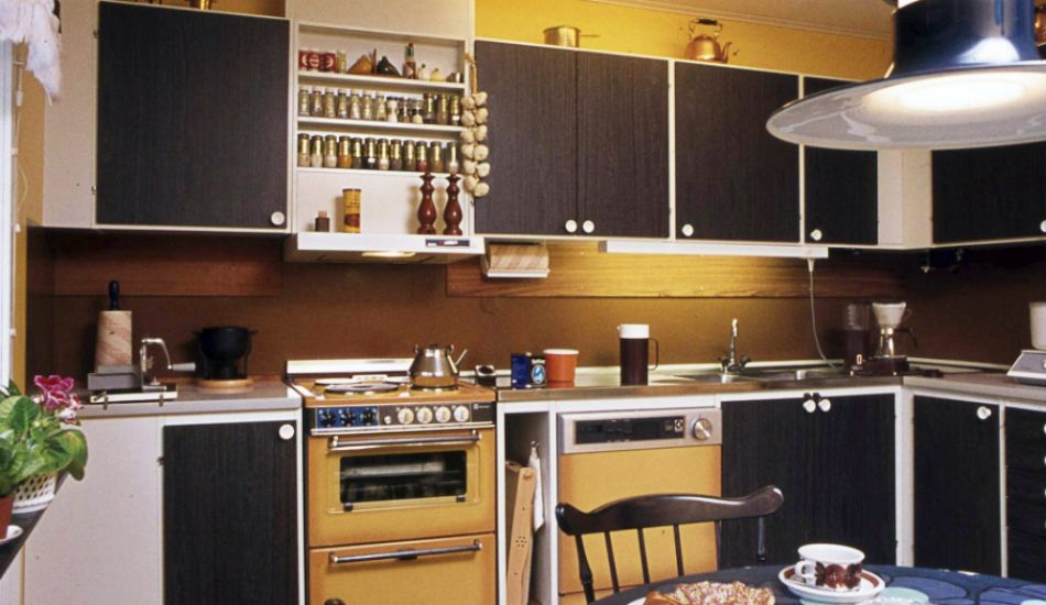 Kök med mörka skåpsluckor och gula inslag, inklusive en gul spis och vägghängda kryddhyllor. Bordet framme i bilden är dukat med kakor och kaffe