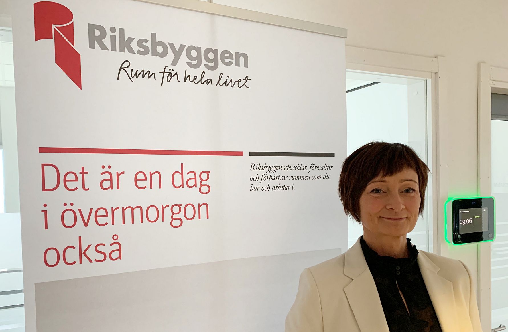 Person i ljus kavaj står bredvid en skylt med texten "Riksbyggen - Rum för hela livet" och "Det är en dag i övermorgon också