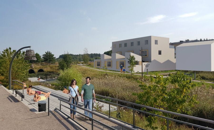 Två personer går på en gångväg vid ett naturskönt område med gräs, träd och vattendrag; i bakgrunden syns modern bebyggelse och solbadande människor