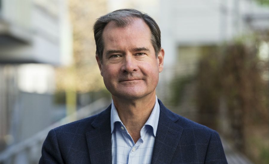 Man i kostymjacka och skjorta står utomhus med en suddig bakgrund av byggnader och vegetation i dagsljus