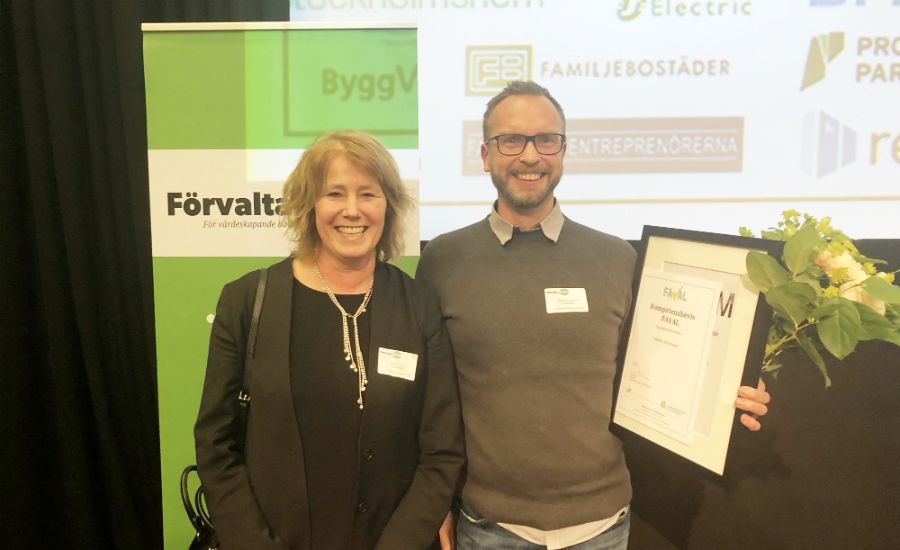 Två personer poserar framför en grön bakgrund med logotyper, där en av dem håller en inramad utmärkelse och en bukett blommor