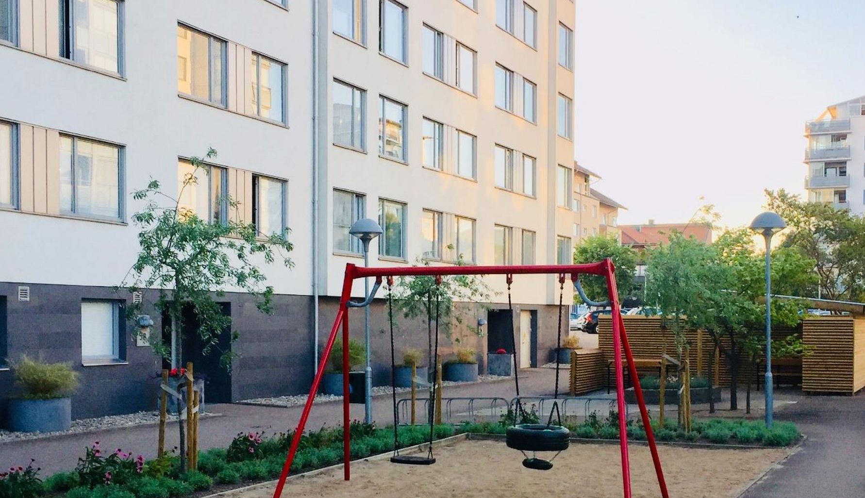 Lekplats med en röd gungställning och sandyta framför ett flerfamiljshus med vita fasader och flera fönster