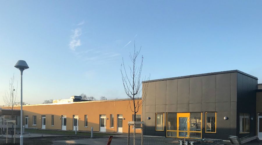 En modern byggnad med grå och gul fasad, stora fönster och en ingång. Området framför byggnaden har belysningsstolpar, träd och trottoar