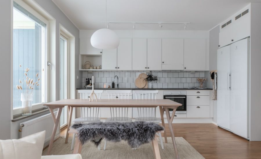 Ljus och modern köksinredning med träbord, vita stolar och pälsöverdrag. Vita skåpluckor, grått kakel och stora fönster släpper in naturligt ljus