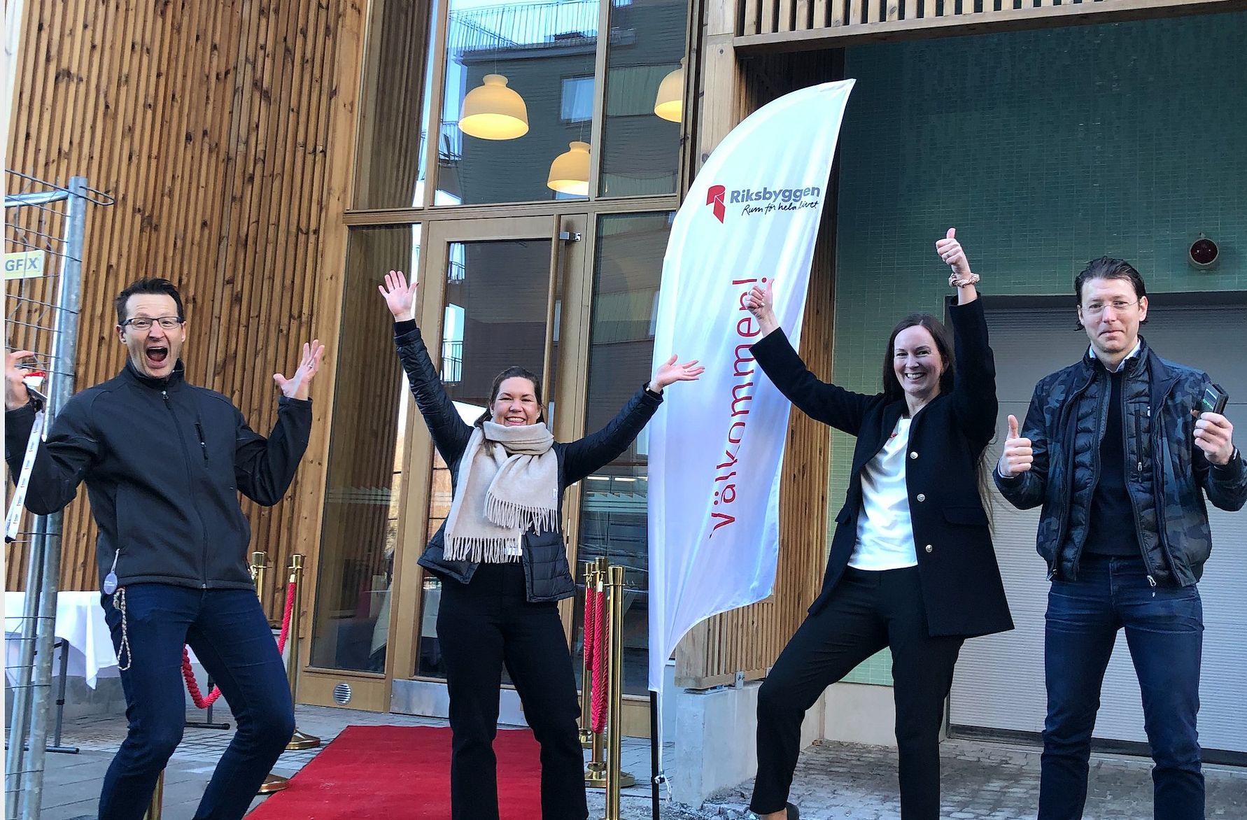 Grupp av fyra personer poserar glada framför en byggnad med en röd matta, ett välkomstflagg och glasdörrar i bakgrunden