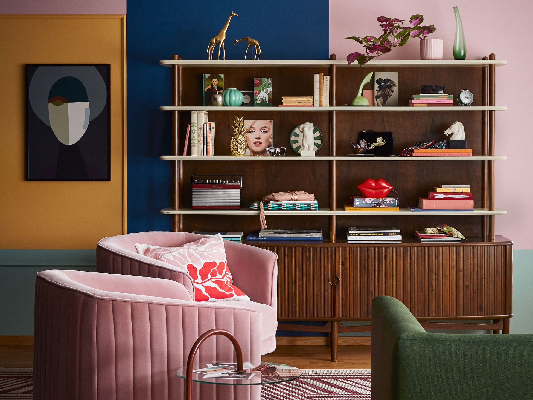 Vardagsrum med färgglada väggar, en hylla fylld med böcker, dekorativa föremål och växter, samt möbler inklusive en rosa fåtölj, ett grönt soffhörn och ett runt glasbord