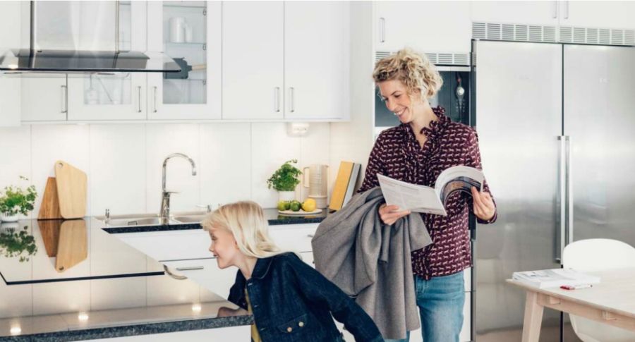 Kvinna i en modern ljus köksmiljö med rostfria vitvaror, håller en jacka och ett magasin medan ett barn lutar sig över köksbänken