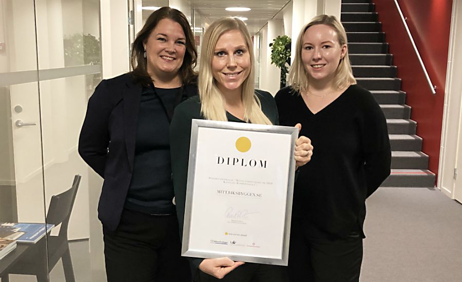 Tre personer står inomhus i en kontorsmiljö, där en person håller upp ett diplom inskrivet med texten "DIPL0M