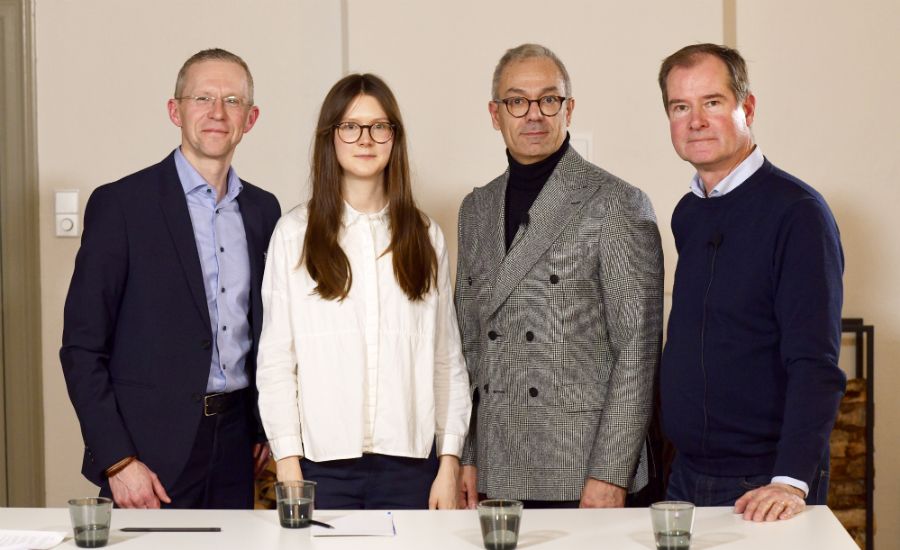 Fyra personer står vid ett bord med glas och dokument, klädda i formella kläder. Bakgrunden är ljus med enkla väggar