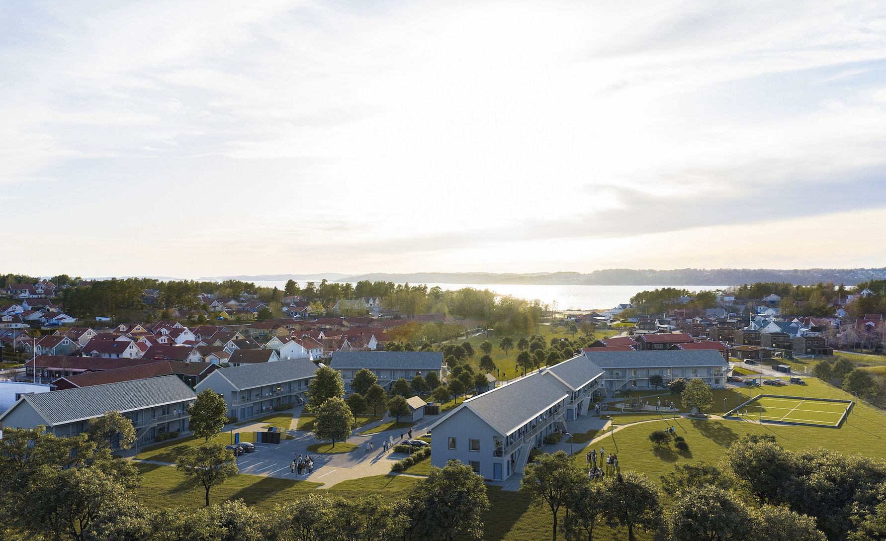 Flygvy över ett bostadsområde med gräsmattor, träd och byggnader, omgiven av hav och skog i bakgrunden under solnedgång