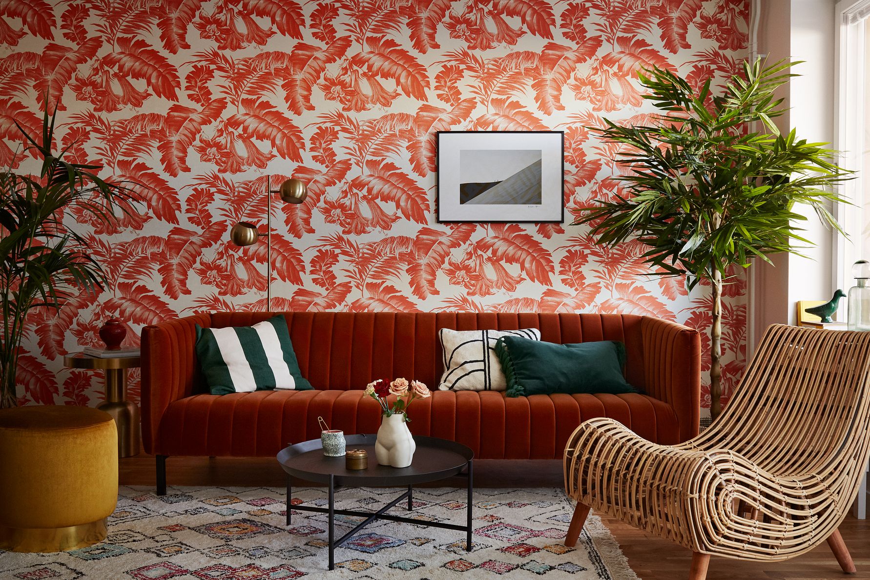 Vardagsrum med orange-röd bladmönstrad tapet, röd sammetssoffa, korgstol, gröna växter och ett soffbord med blommor