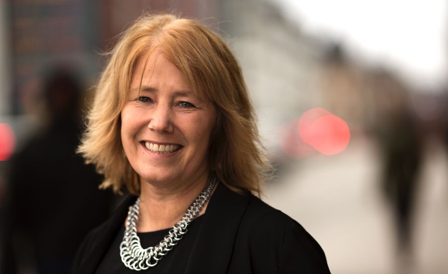 Kvinna med blont hår, klädd i svart outfit och ett silverfärgat halsband, står utomhus med en suddig stadsbakgrund