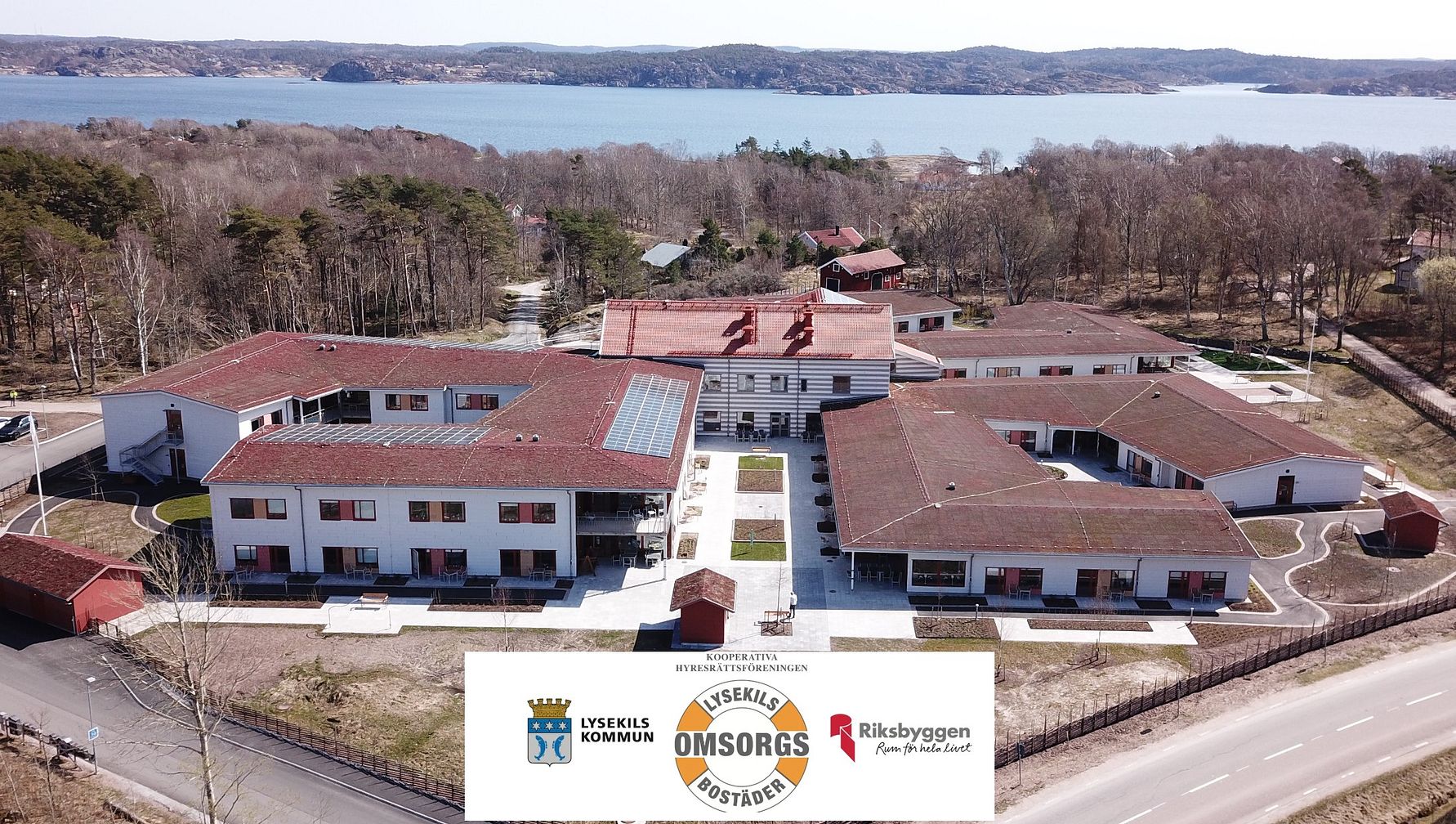 Flygbild av en modern byggnad med röda tak, omgiven av träd och nära en sjö i bakgrunden. Logotyper för olika organisationer syns längst ner i bilden