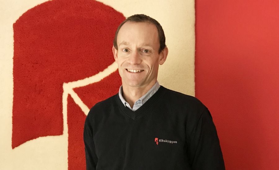 Person i mörk tröja med en logotyp, står framför en vägg med ett stort rött och vitt motiv