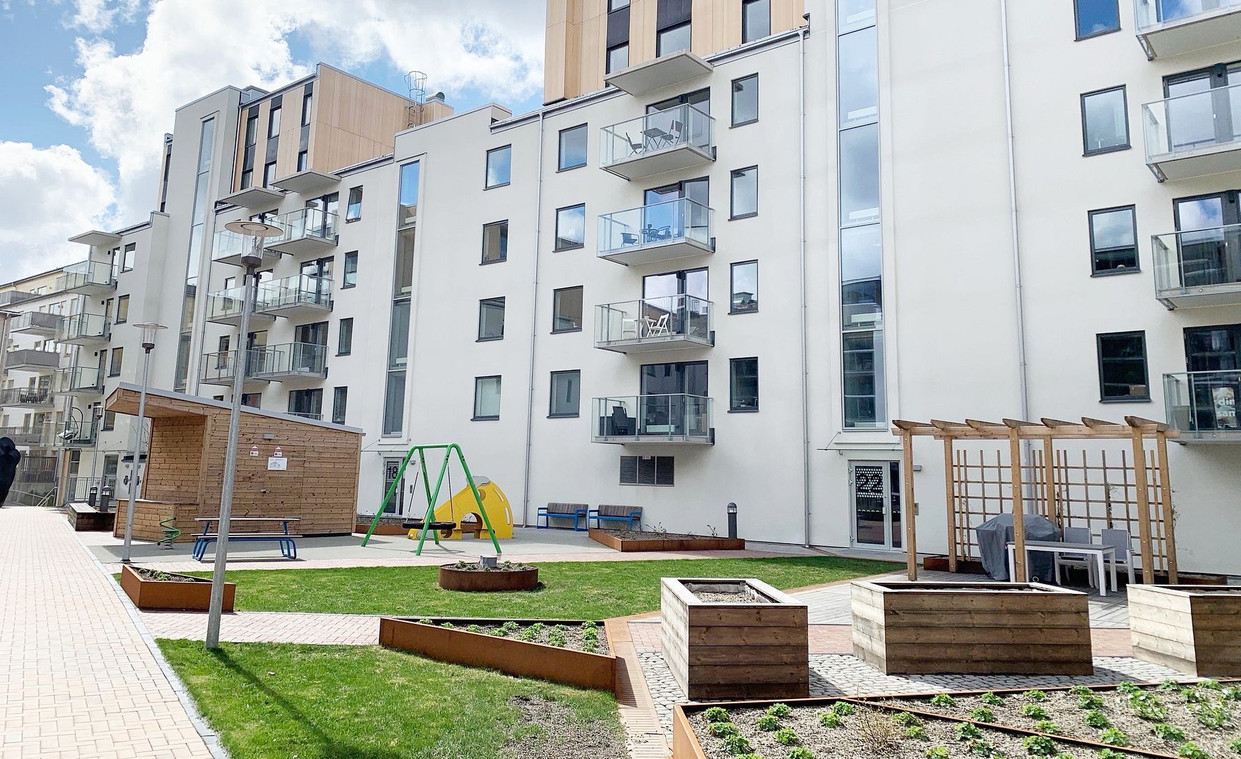 Moderna lägenhetsbyggnader med vit fasad, en lekplats med gungor och gräsmatta samt odlingslådor och sittplatser i förgrunden