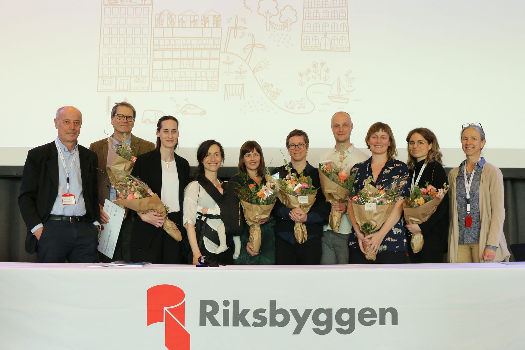 En grupp människor står på en scen framför en vit bakgrund med Riksbyggens logotyp och håller blombuketter