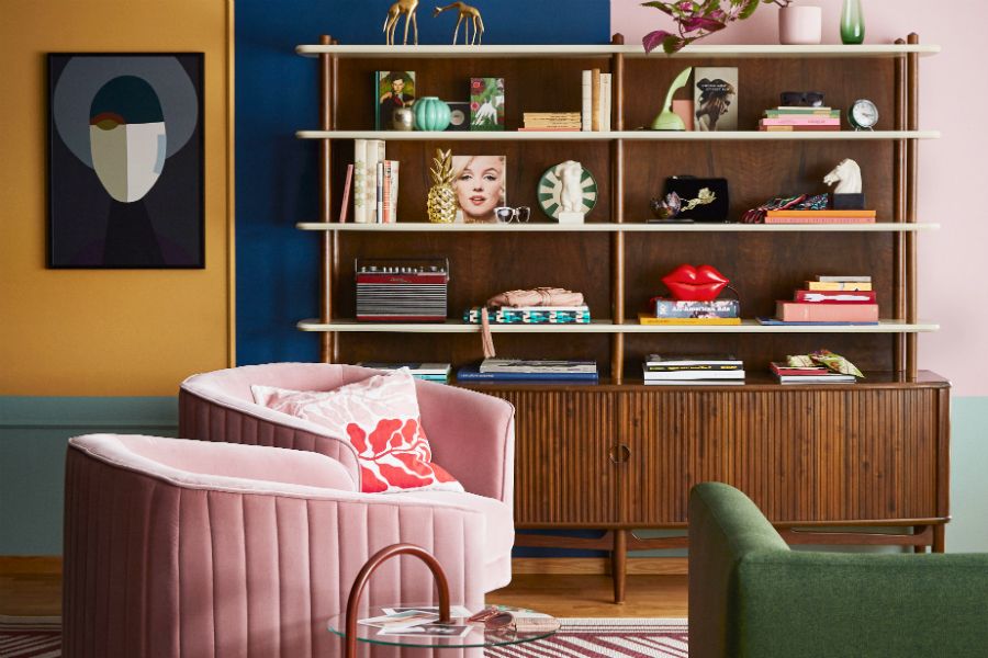Ett vardagsrum med en hylla fylld med böcker, prydnadsföremål och växter. Framför hyllan står en rosa fåtölj, ett grönt soffhörn och ett glasbord