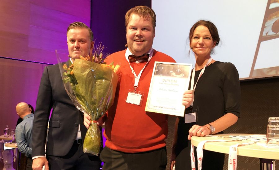 Tre personer står inomhus vid ett bord, där en av dem håller ett diplom och en blombukett. Bakgrunden visar en scen och en projektion på väggen