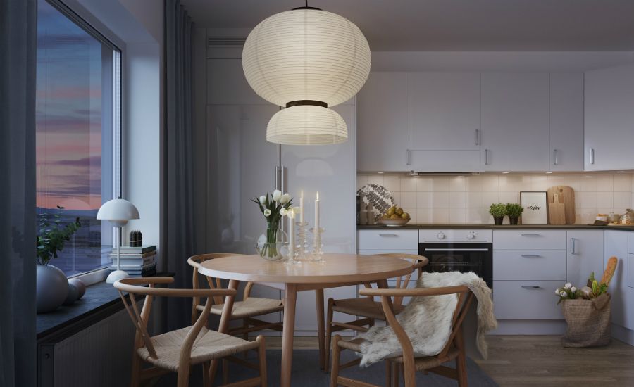 Matbord med trästolar och en rund taklampa i ett modernt kök med vita skåp, bänkskiva och dekorativa detaljer. Fönster med kvällsutsikt och gardiner