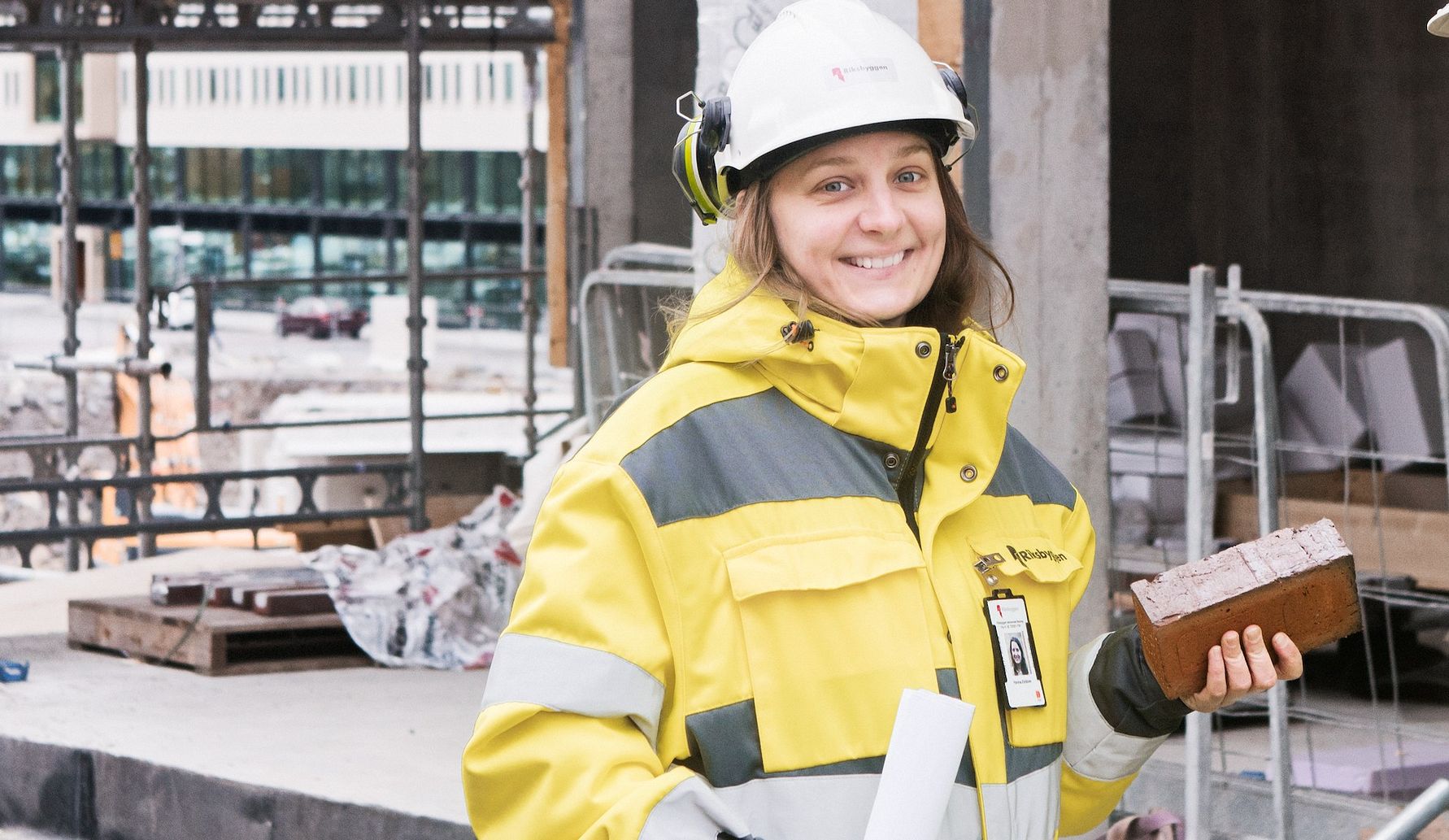 Person i gul arbetsjacka och hjälm som står på en byggarbetsplats och håller i en tegelsten och ett papper