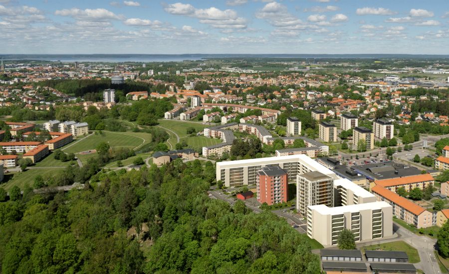 Flygfoto över ett bostadsområde med flerfamiljshus, gröna parkområden och stadsmiljö i bakgrunden under en klarblå himmel med några moln