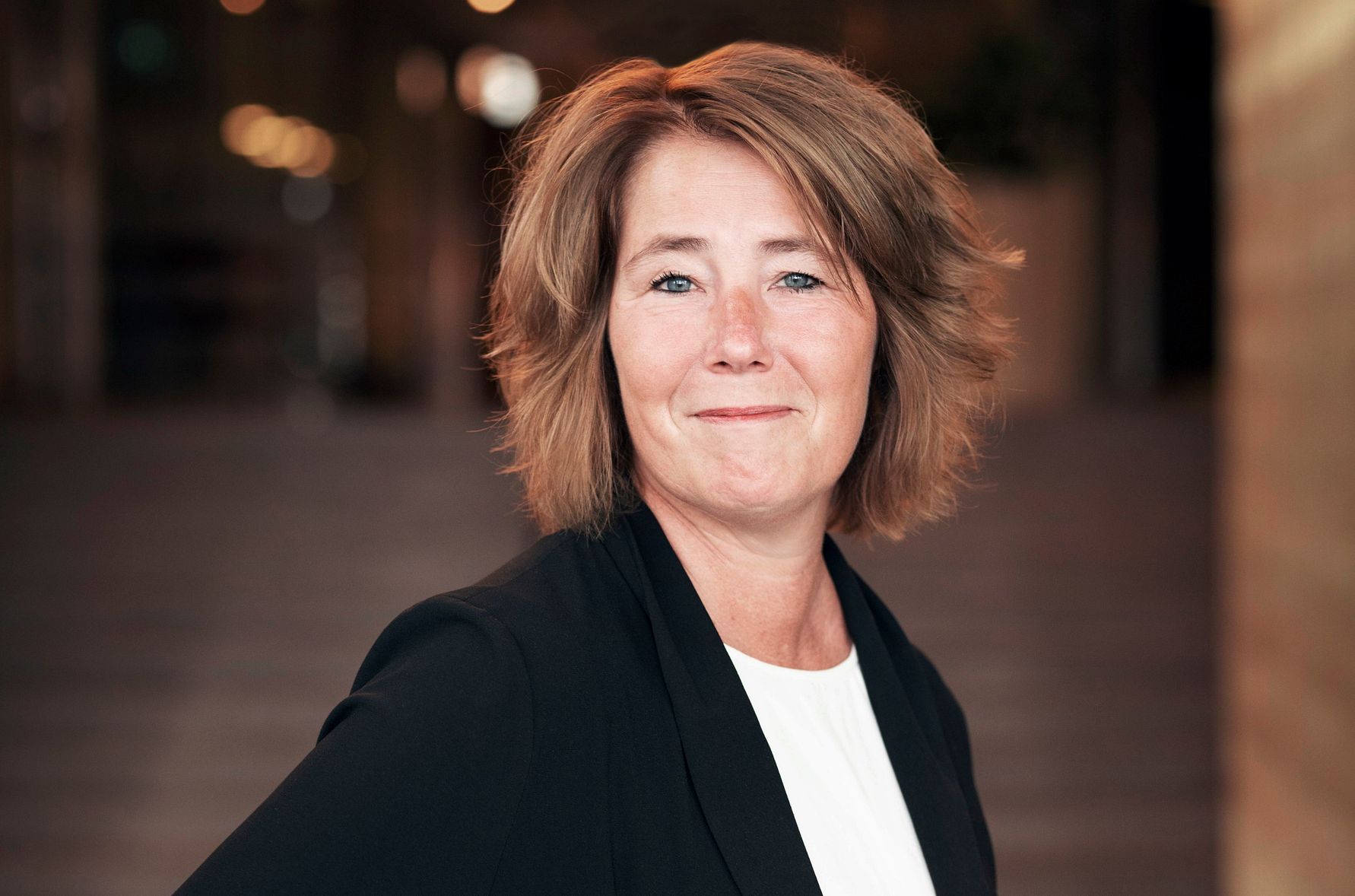 Kvinna i mörk kavaj och vit topp, med axellångt blont hår, stående framför en suddig bakgrund av en trappa inomhus