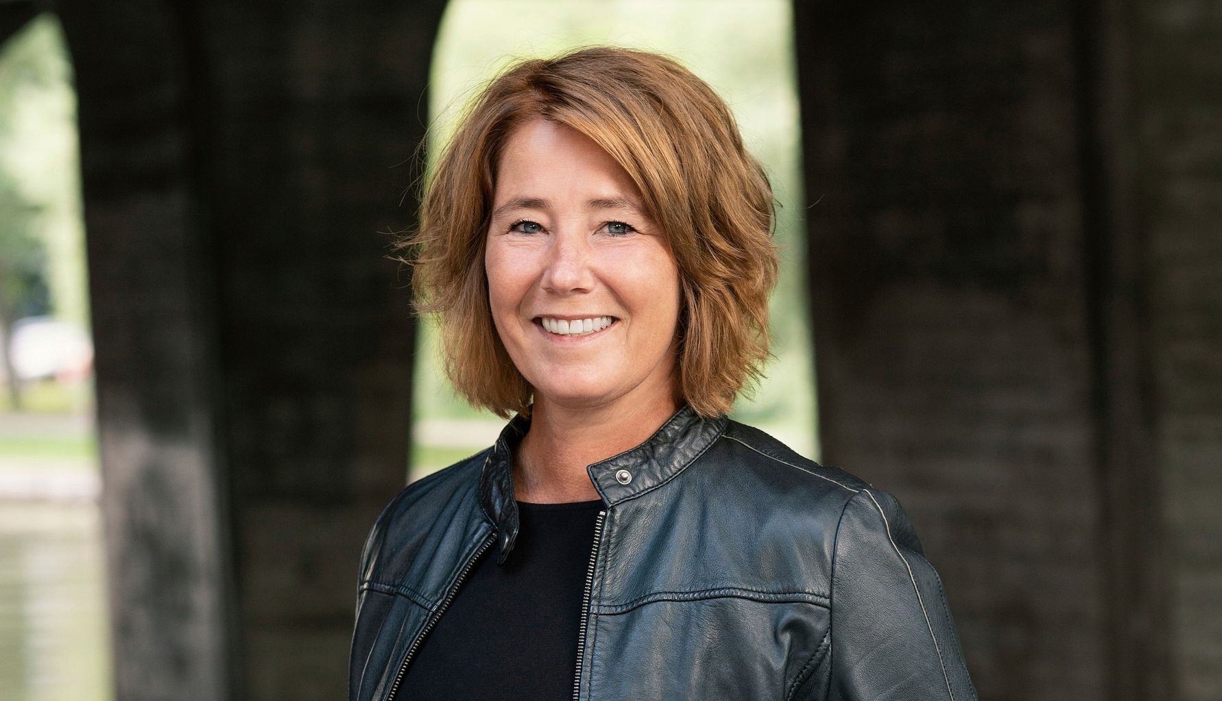 Person i svart läderjacka och svart tröja står framför en bakgrund med bågar av sten och grönska i bakgrunden