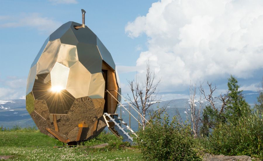 Ett äggformat, guldglänsande bastuhus med geometrisk design, placerat i en grönskande natur med berg och moln i bakgrunden