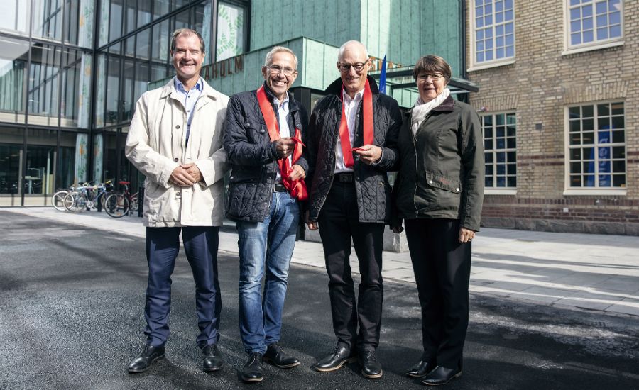 Fyra personer står utomhus framför en modern byggnad och en tegelvägg, med cyklar synliga i bakgrunden. Två av dem bär röda halsdukar