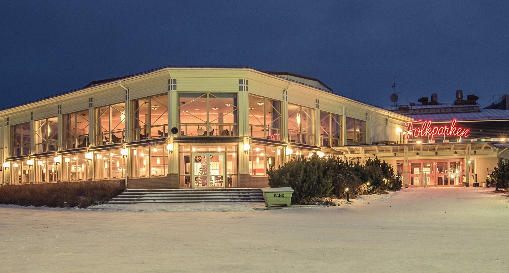 Byggnad med glasfasad och neonskylt som lyser "Folkparken", omgiven av snötäckt mark under kvällstid