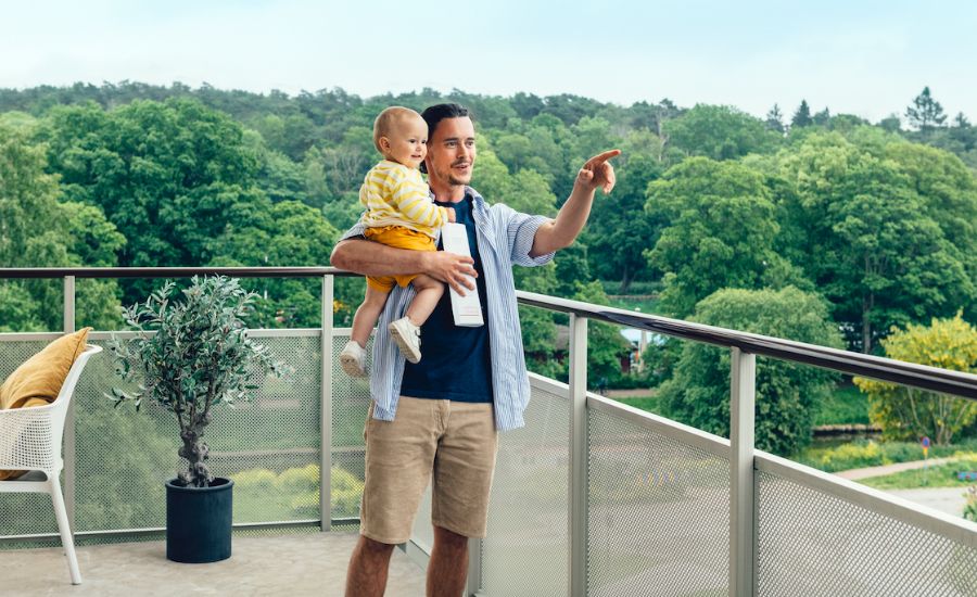En man på en balkong håller ett litet barn i famnen och pekar ut mot en grönskande utsikt
