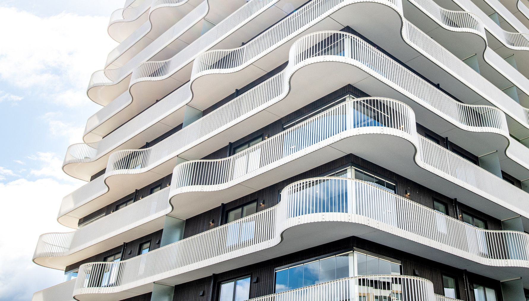 Modern byggnad med flera våningsplan, vit vågformad balkongdesign och mörka fasadelement, mot en bakgrund av blå himmel och moln