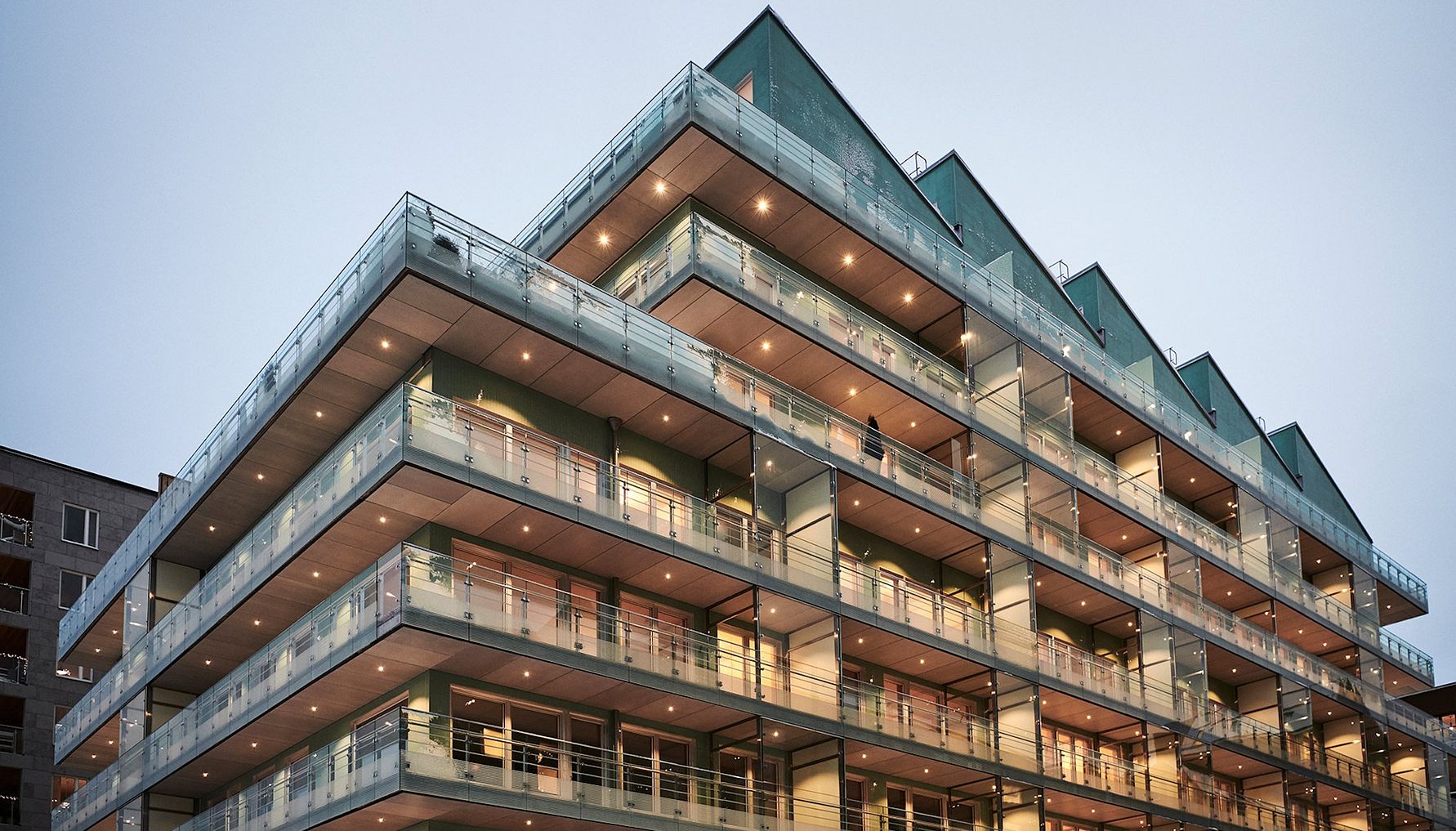 Modernt flervåningshus med glasräcken och belysning, fotograferat från marknivå mot en molnig himmel