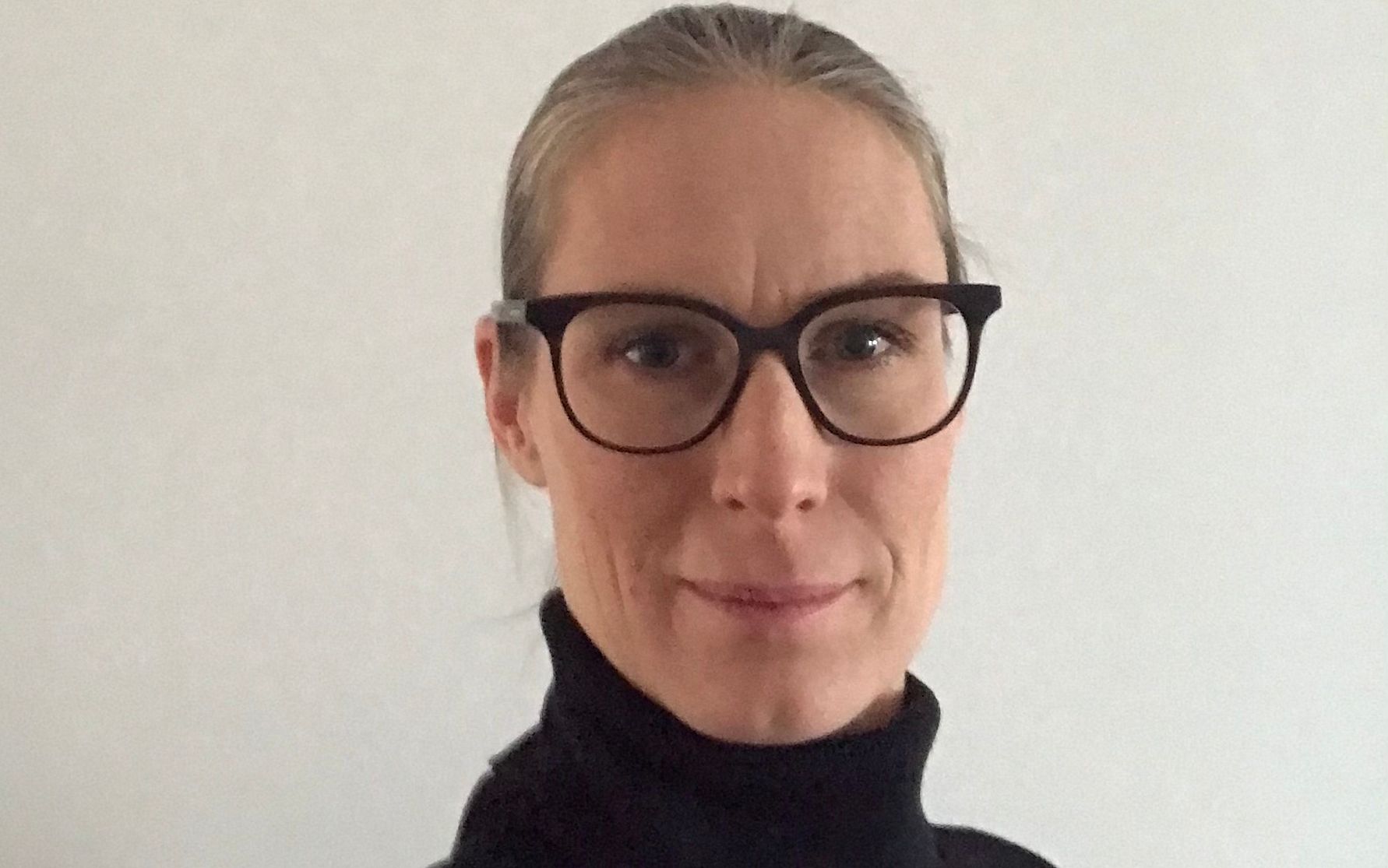 En person med blont hår uppsatt i en låg frisyr, som bär svarta glasögon och en svart polotröja, framför en vit bakgrund