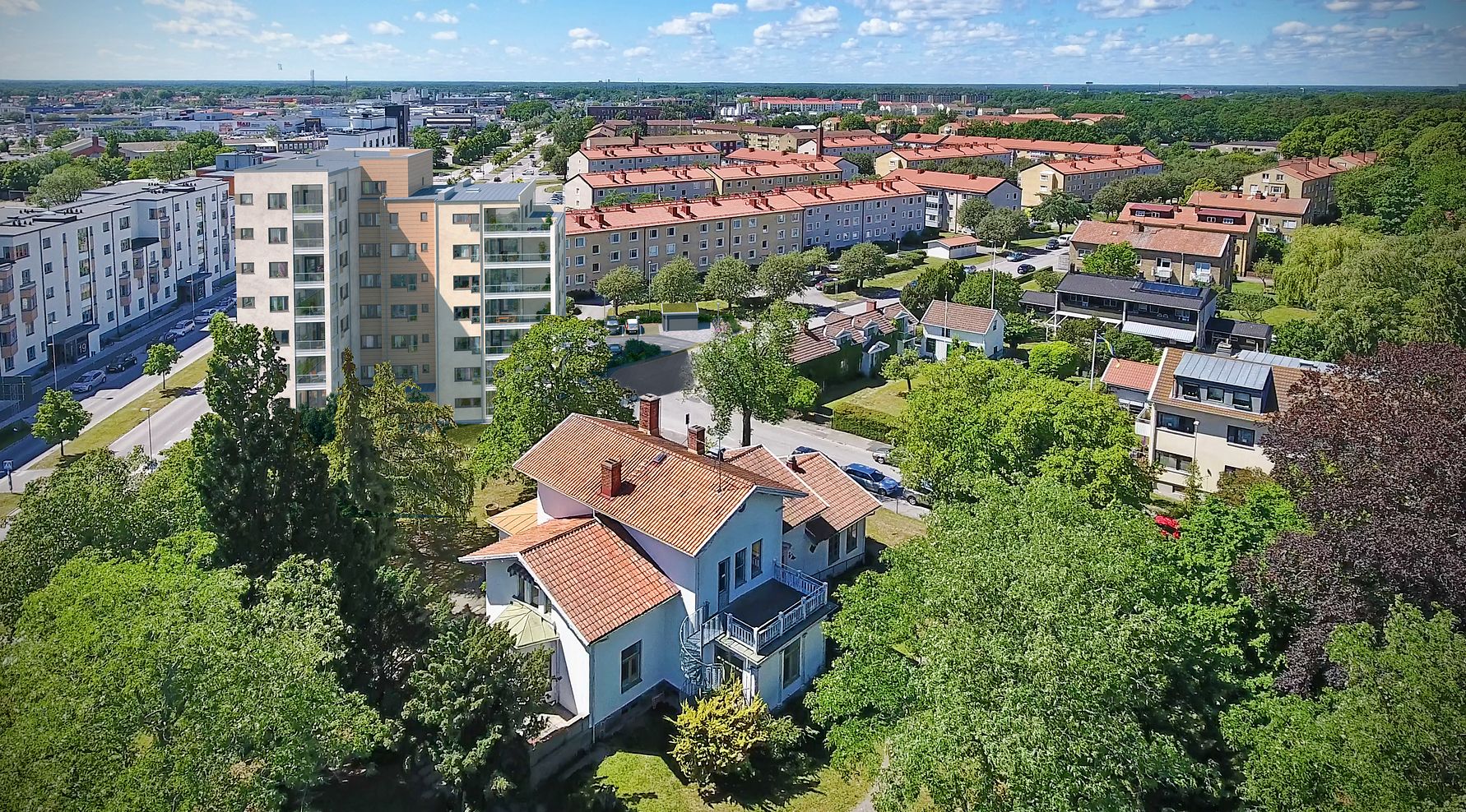 Luftbild av ett bostadsområde med villor, flerfamiljshus och grönområden under en klar blå himmel