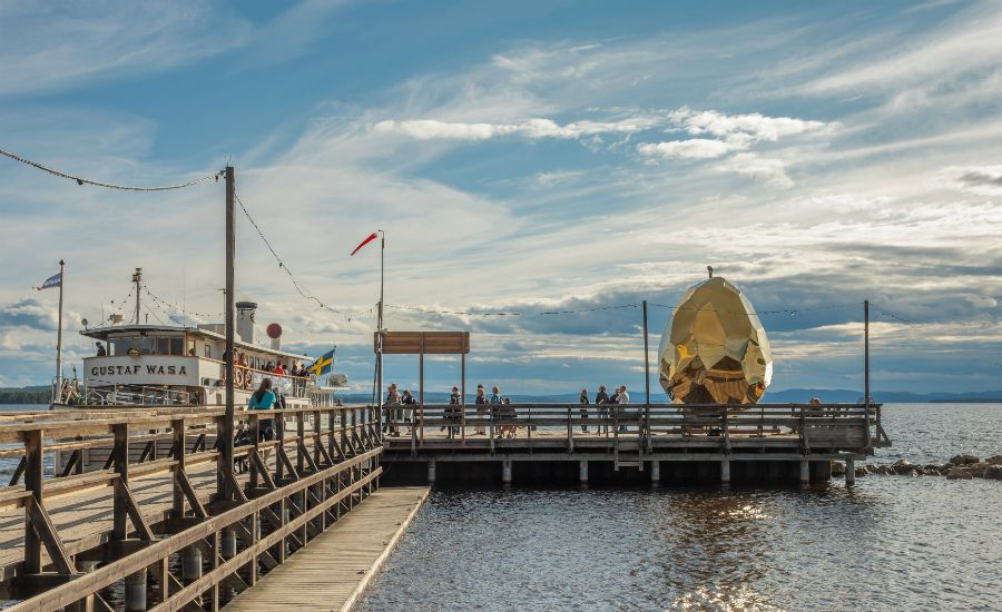 Brygga med gångvägar vid en sjö, med båten "Gustaf Wasa" till vänster och en guldfärgad konstinstallation till höger under en blå himmel med moln