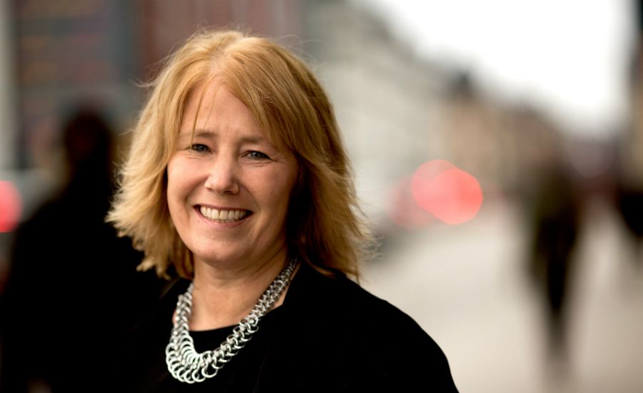 En person med blont hår klädd i svart och ett halsband av silverkedjor, i en stadsmiljö med suddig bakgrund