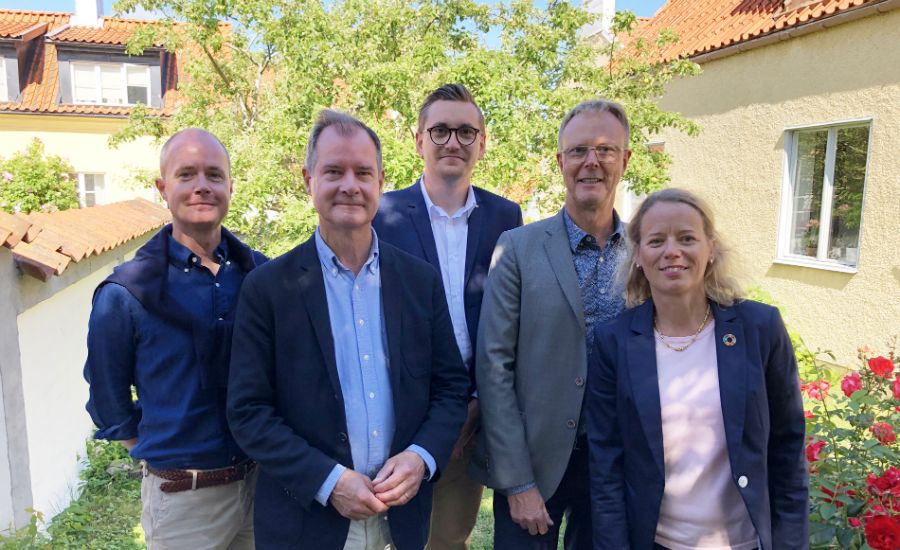En grupp människor står utomhus framför hus med gula fasader och tegeltak, omgivna av gröna träd och röda blommor