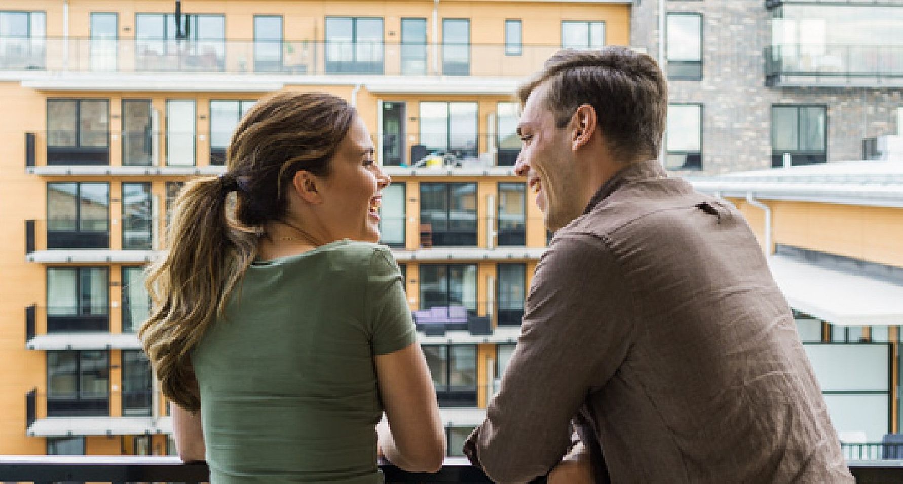 Två personer står på en balkong och tittar ut över ett modernt bostadsområde med orange fasader och stora fönster