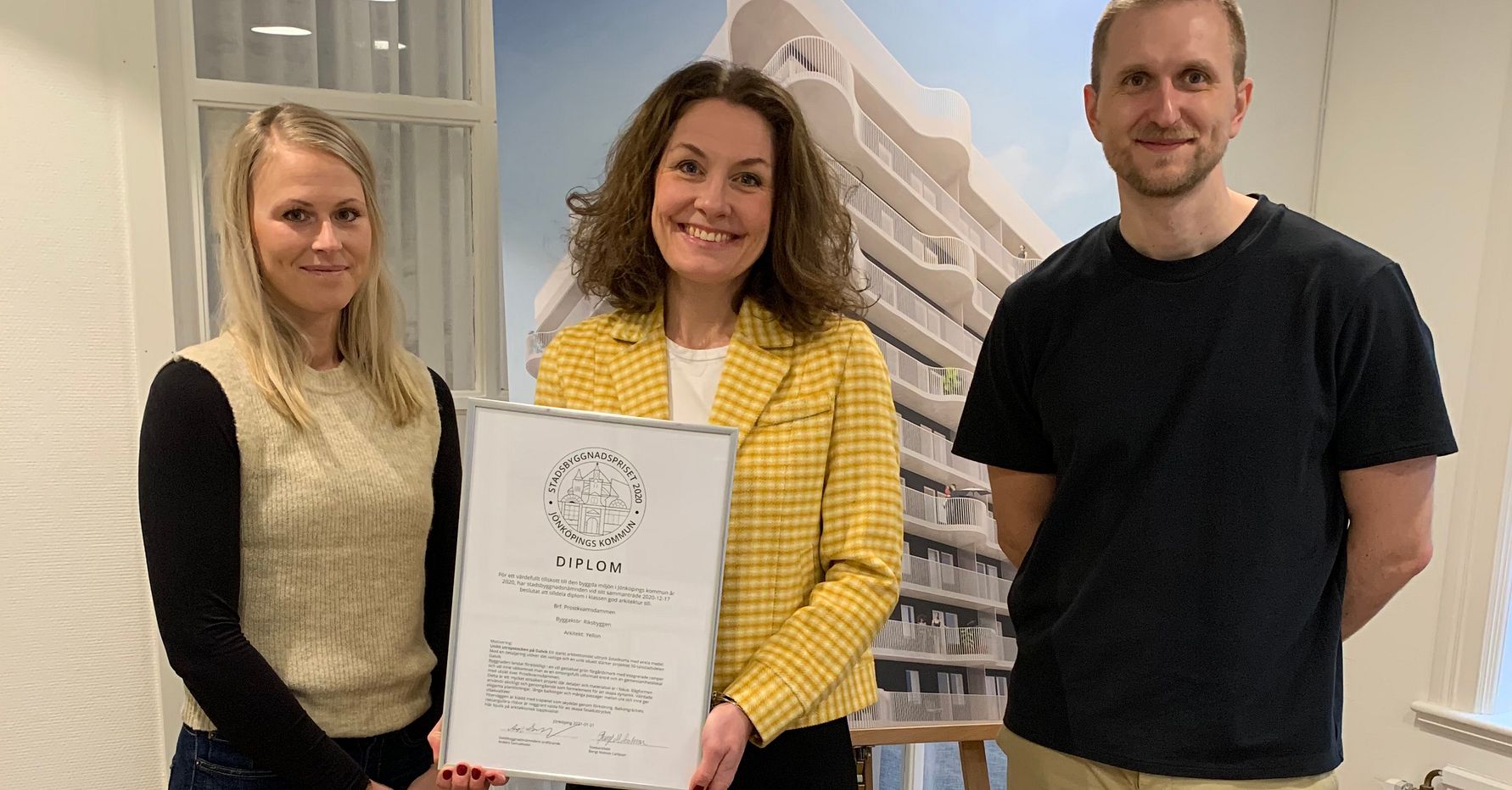 Tre personer står framför ett fönster och en vägg med en stor affisch av en modern byggnad, där den mittersta personen håller ett diplom