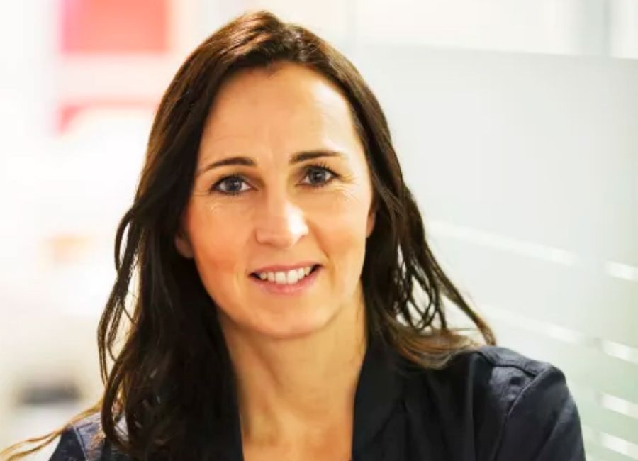 Person med mörkt hår och mörkblå skjorta, sittande framför en ljus bakgrund med glas och suddiga detaljer i röda och vita nyanser