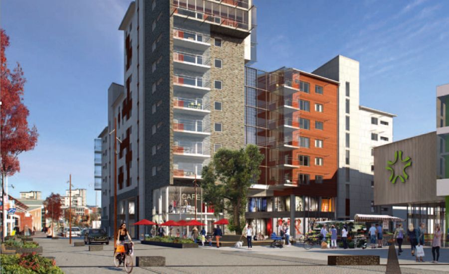 Modern stadsbild med höga bostadshus, kaféer och butiker, omgiven av gångväg och cyklister, med träd och klar blå himmel i bakgrunden